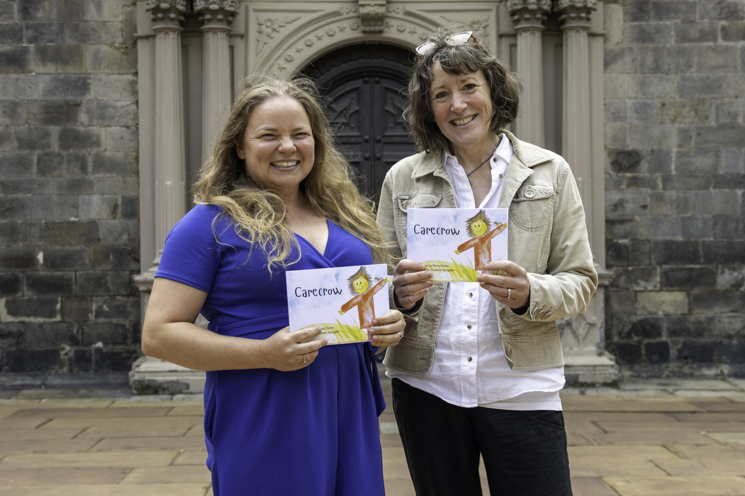 two people standing outside, smiling, and holding a book